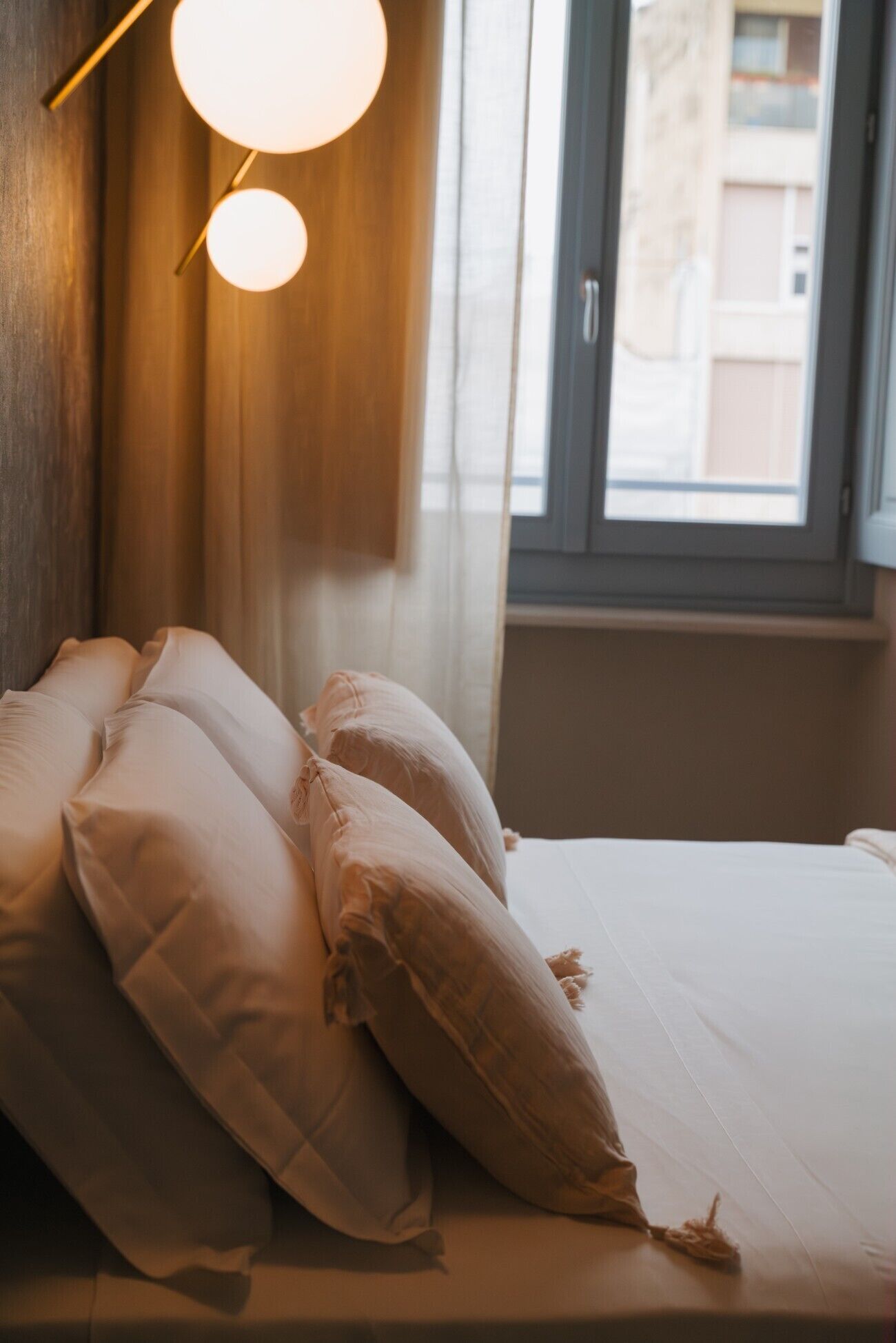 Una cama con sábanas y almohadas blancas, cabecera de madera y ventana con vistas a edificios.