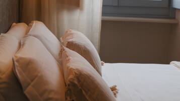 Una cama con sábanas y almohadas blancas, cabecera de madera y ventana con vistas a edificios.