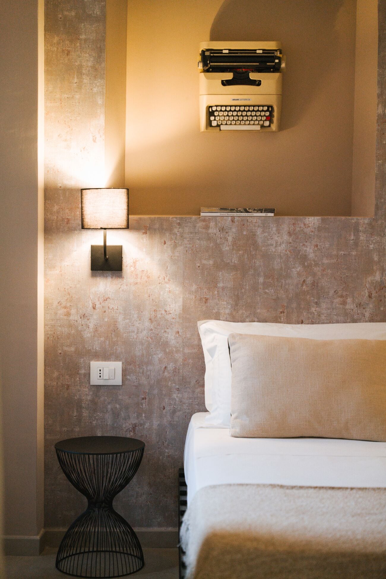 Un dormitorio con cama, mesita de noche, lámpara, estante con libros y pared texturizada.