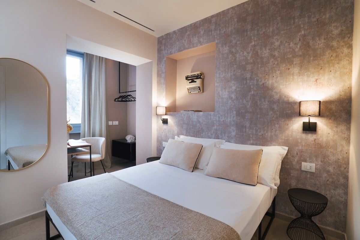 A modern hotel room with a large bed, a round mirror, a desk, and a chair.