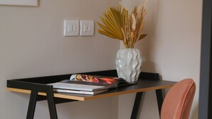Ein Schreibtisch mit einem Laptop, einem Buch und einer Vase mit Trockenpflanzen. Ein Stuhl mit Kissen und eine Steckdose sind ebenfalls vorhanden.