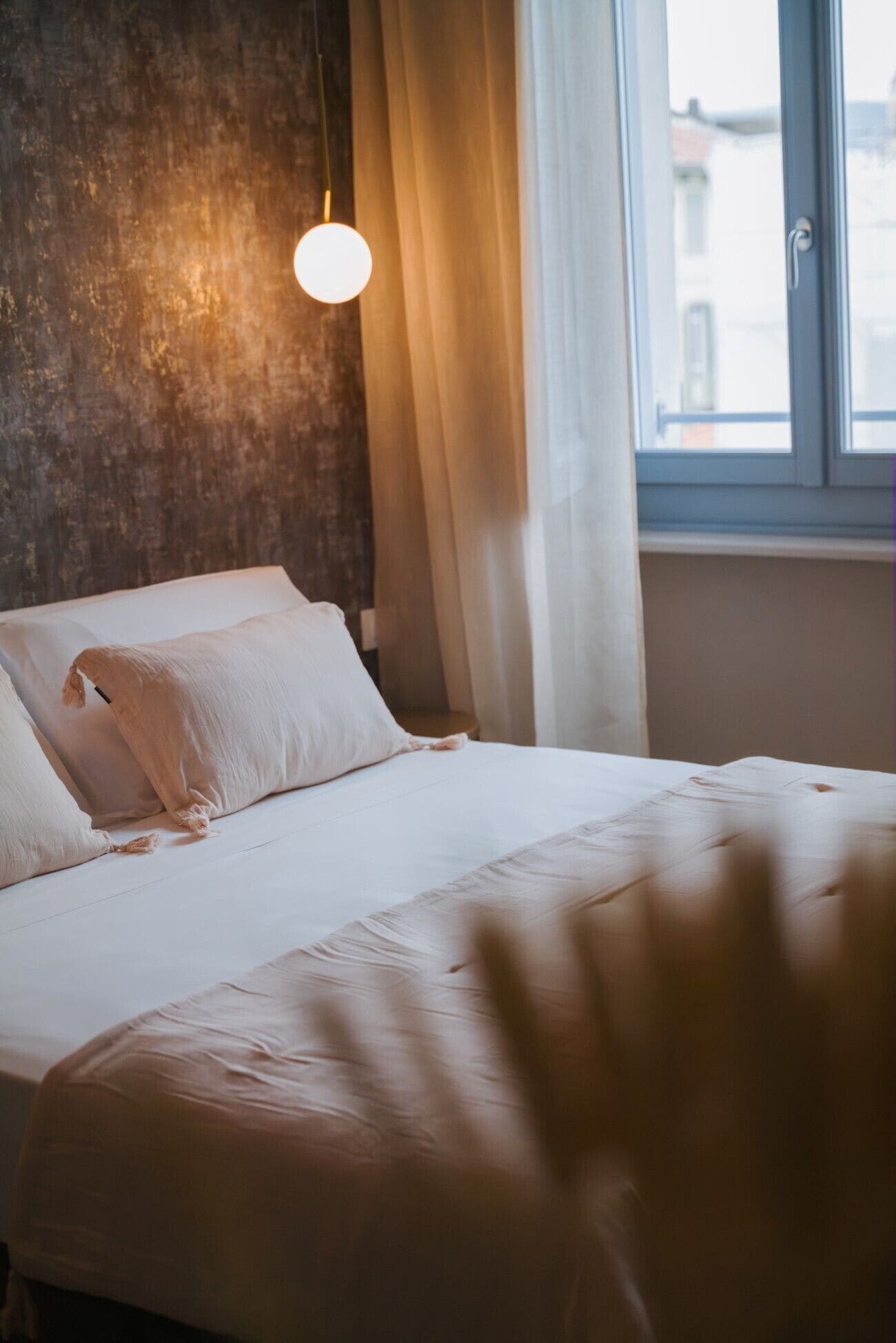 Een bed met witte lakens, een hanglamp en een raam met gordijnen.