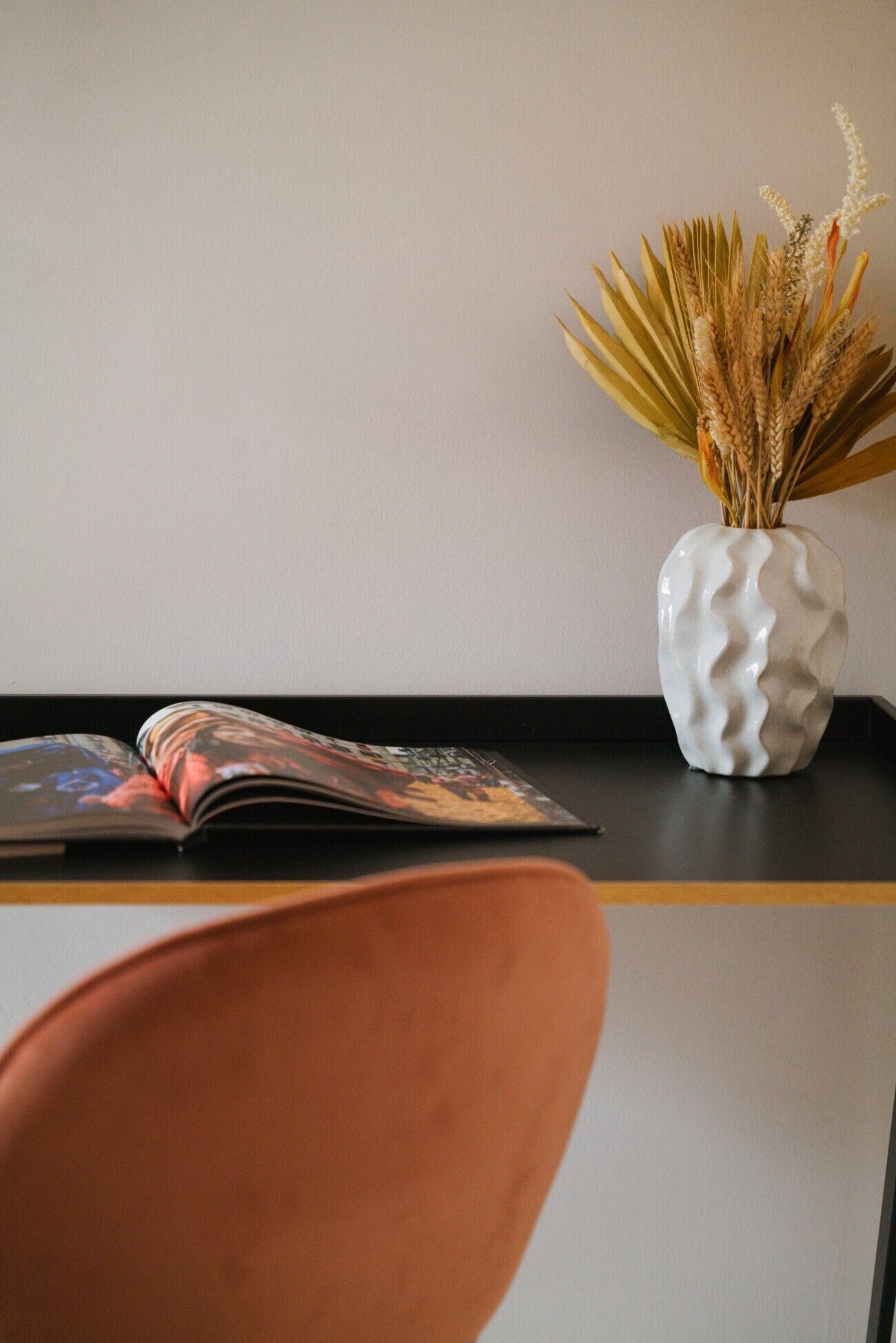 A magazine on a black table, a white vase with dried plants, and a brown chair.