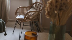 Ein Zimmer mit einem Korbstuhl, einem kleinen Tisch mit Kerze und einer Vase mit Trockenpflanzen.