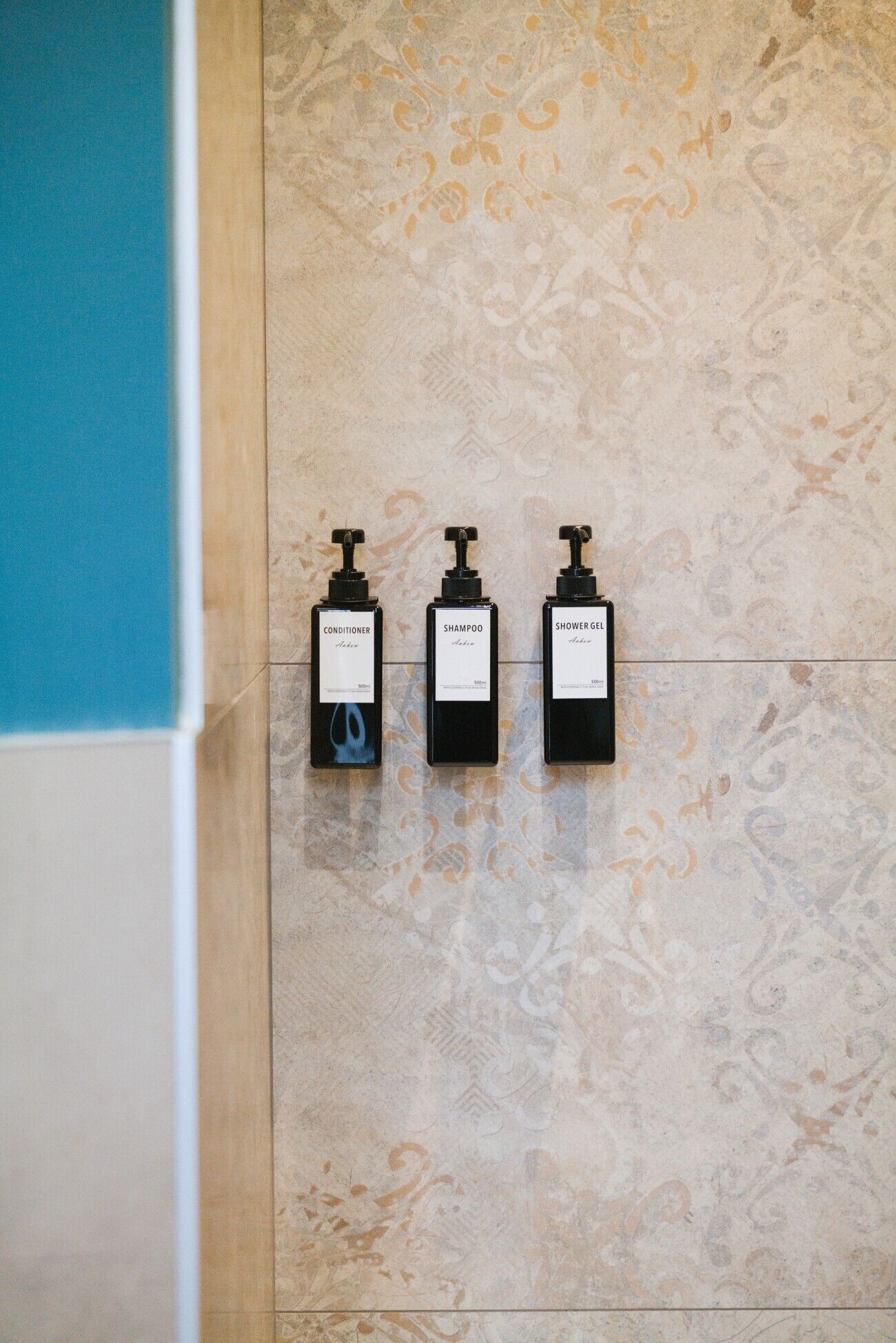 Three black bottles labeled conditioner, shampoo, and shower gel on a tiled wall.