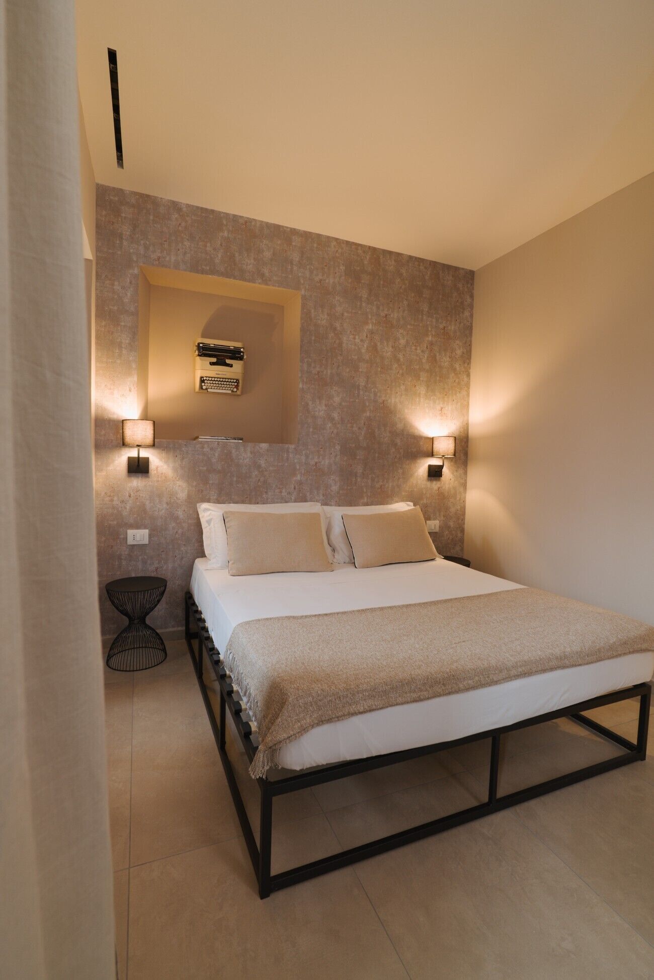 Un dormitorio moderno con una cama, dos lámparas de noche, una mesita redonda y un estante de pared con libros.