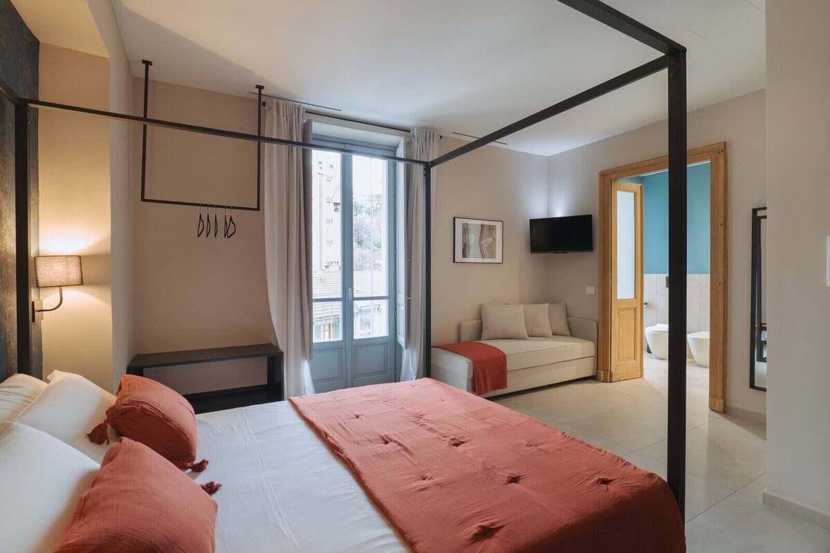 A modern hotel room with a large bed, a sofa, a TV, and a bathroom visible through an open door.