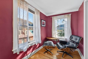 Una habitación con dos ventanas, un sillón de cuero negro, una mesita de madera y un cuadro en la pared.