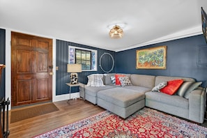 Una sala de estar con un sofá modular gris, una puerta de madera, una pared azul decorativa, un tapete estampado y una lámpara en el techo.