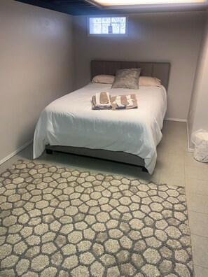 Un lit bien fait, agrémenté d’un tapis à motifs, une petite fenêtre et un mur sobre.