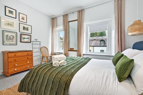 Une chambre à coucher avec un lit, une literie verte et blanche, une commode en bois, une chaise et des tableaux encadrés accrochés au mur.