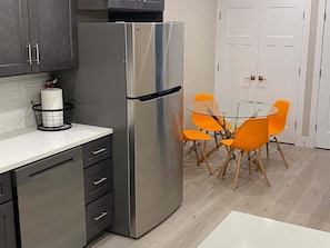 A kitchen with a stainless steel refrigerator, a white countertop, a glass dining table with orange chairs, and a white door in the background.