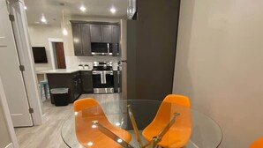 A modern kitchen with dark cabinets, stainless steel appliances, and a glass dining table with orange chairs.