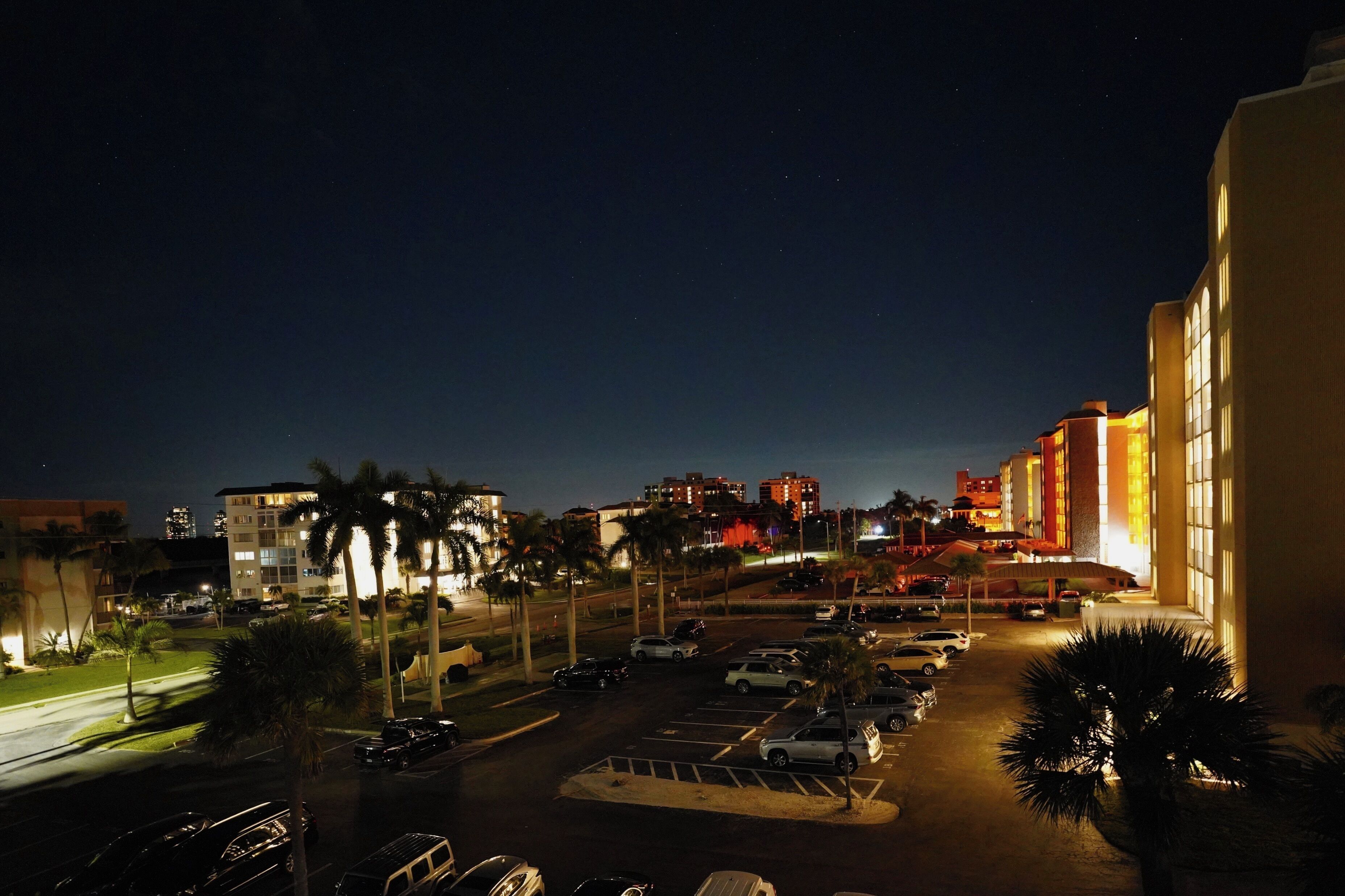A nighttime view of a hotel with a parking lot, palm trees, and illuminated buildings in the background.