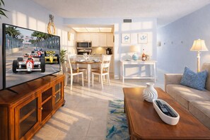 A modern living room with a television displaying a racing car, a wooden cabinet, a coffee table with a remote control, a beige sofa, and a dining area with white chairs and a kitchen in the background. - "Sunset Coast Retreat" – King Bed – Gulf Front – Bonita Springs (Bonita Springs)