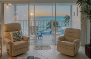 A balcony with two beige armchairs, a small table, and a chair with a blue cushion. The balcony overlooks a pool and the ocean. - "Sunset Coast Retreat" – King Bed – Gulf Front – Bonita Springs (Bonita Springs)