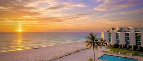 A beachfront hotel with a pool, palm trees, and a sunset view.