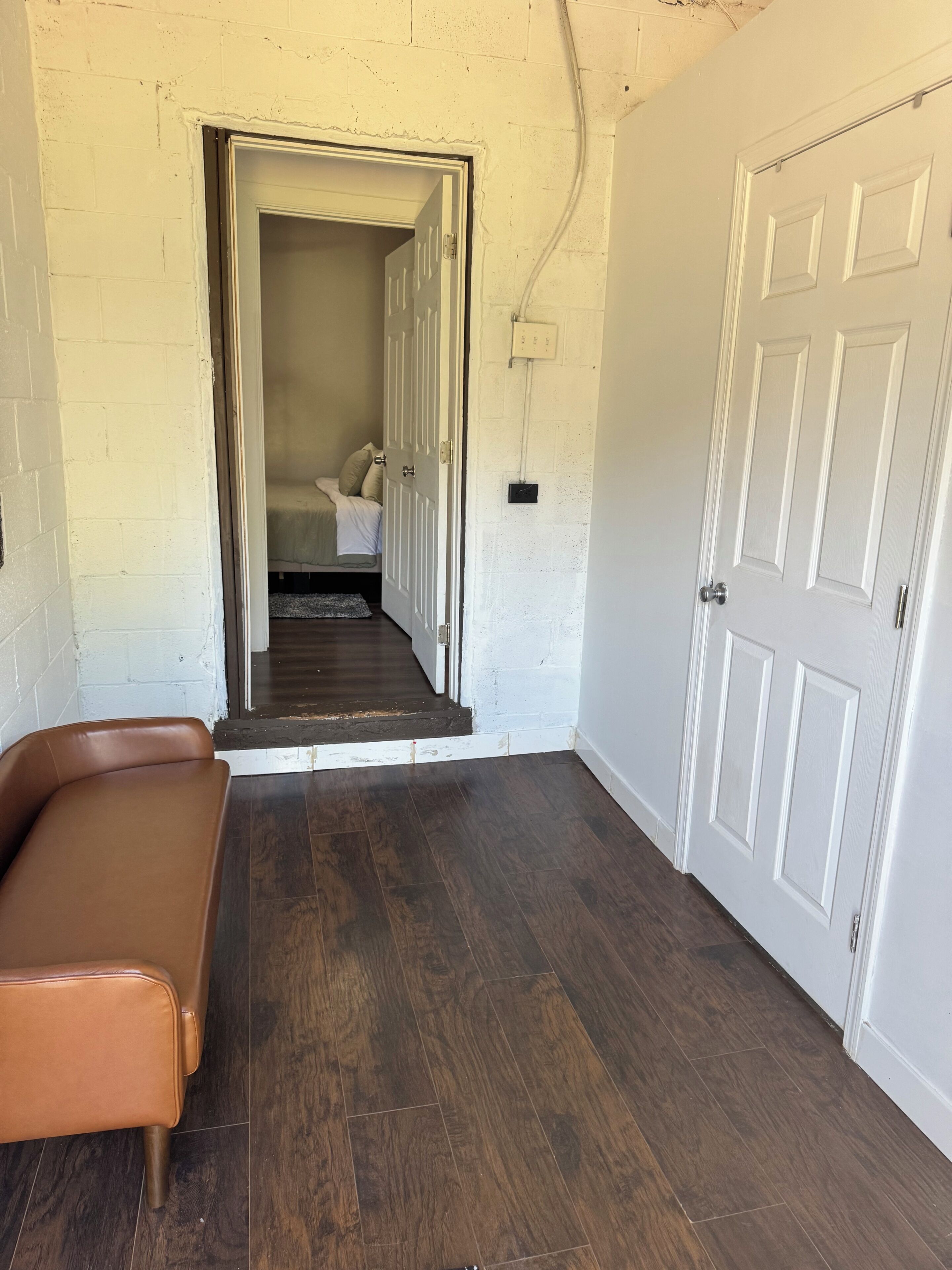 Un couloir avec un banc en cuir marron, une porte blanche et un escalier menant à une chambre à coucher équipée d’un lit et d’une table de chevet.