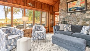 A cozy living room with a stone wall, wooden ceiling, and large windows. There are two patterned armchairs, a blue sofa, and a white ottoman. A painting hangs on the stone wall, and a lamp is on a wooden table. - Private FLX Retreat | 200' Seneca Lakefront | Pool (Geneva)