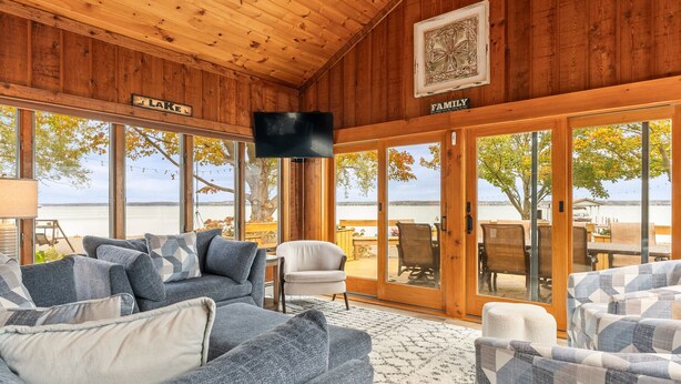 A cozy cabin with a large window overlooking a lake, furnished with blue and white patterned sofas and a flat-screen TV mounted on the ceiling. - Private FLX Retreat | 200' Seneca Lakefront | Pool (Geneva)