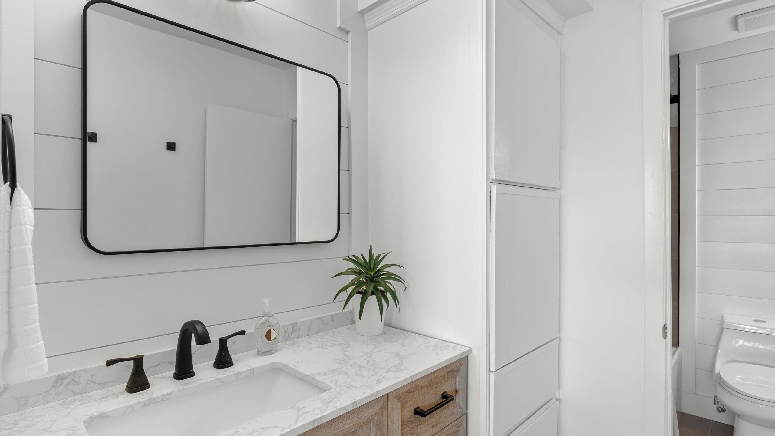 A bathroom with a white sink, black faucets, a mirror, a potted plant, and a toilet.