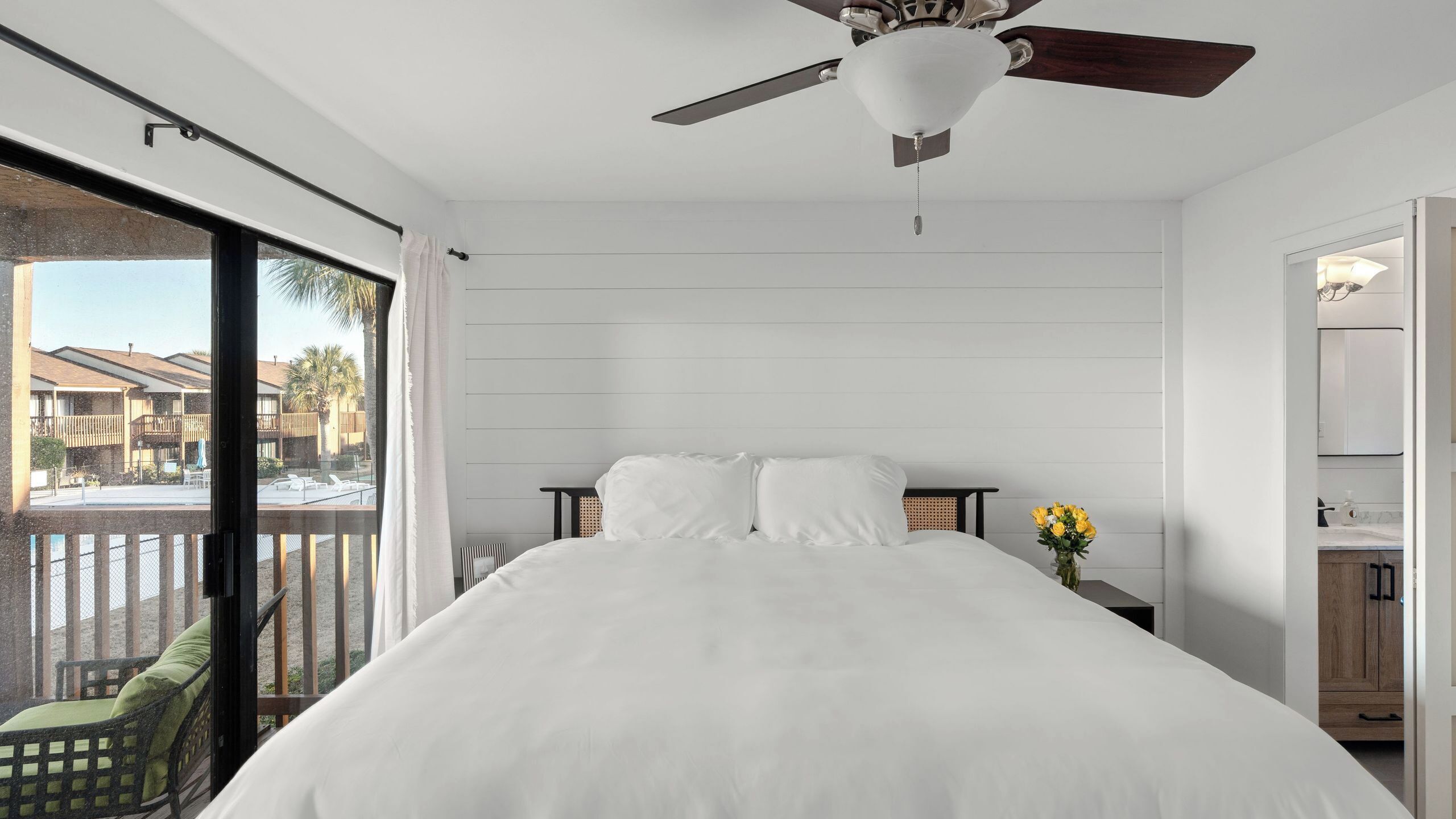 A hotel room with a large bed, a ceiling fan, a view of the outside, a small table with a vase of flowers, and a balcony with a chair.