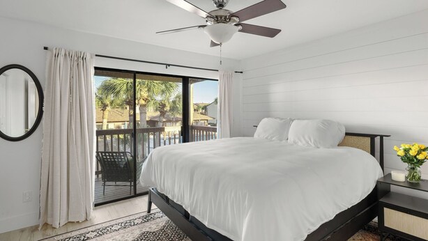 A bedroom with a bed, a ceiling fan, a nightstand with a vase of flowers, and a view of a pool and buildings outside. - Sandbar Serenade by Avantstay Beachside Home + Pool + Tennis Court + Beach Access + Near Pier Park (Panama City Beach)