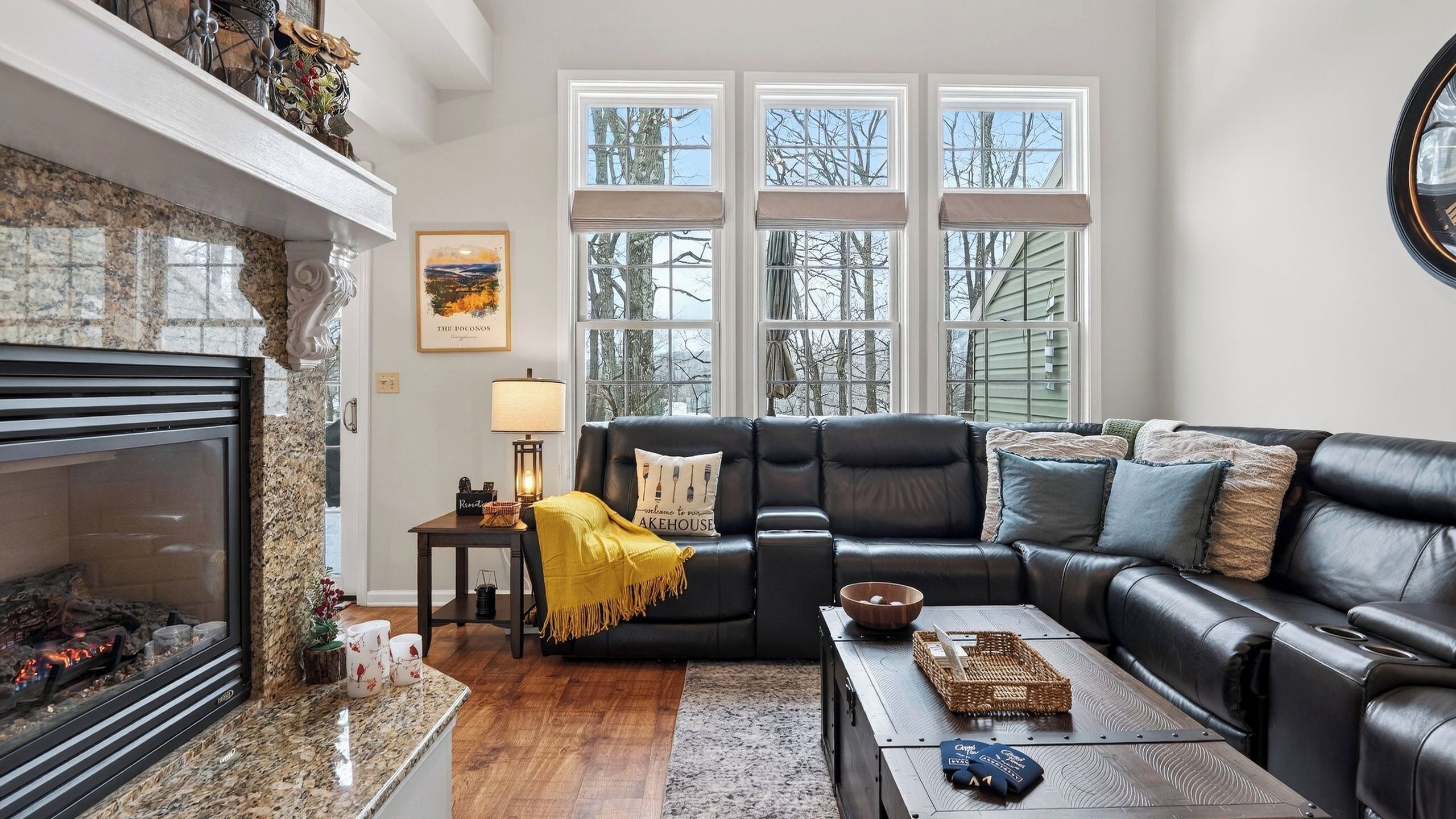 A living room with a fireplace, a black leather sofa, a coffee table, and a view of trees through large windows.