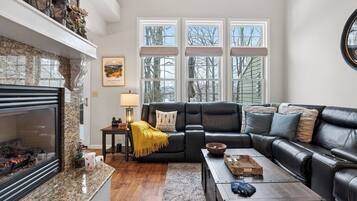 A living room with a fireplace, a black leather sofa, a coffee table, and a view of trees through large windows.