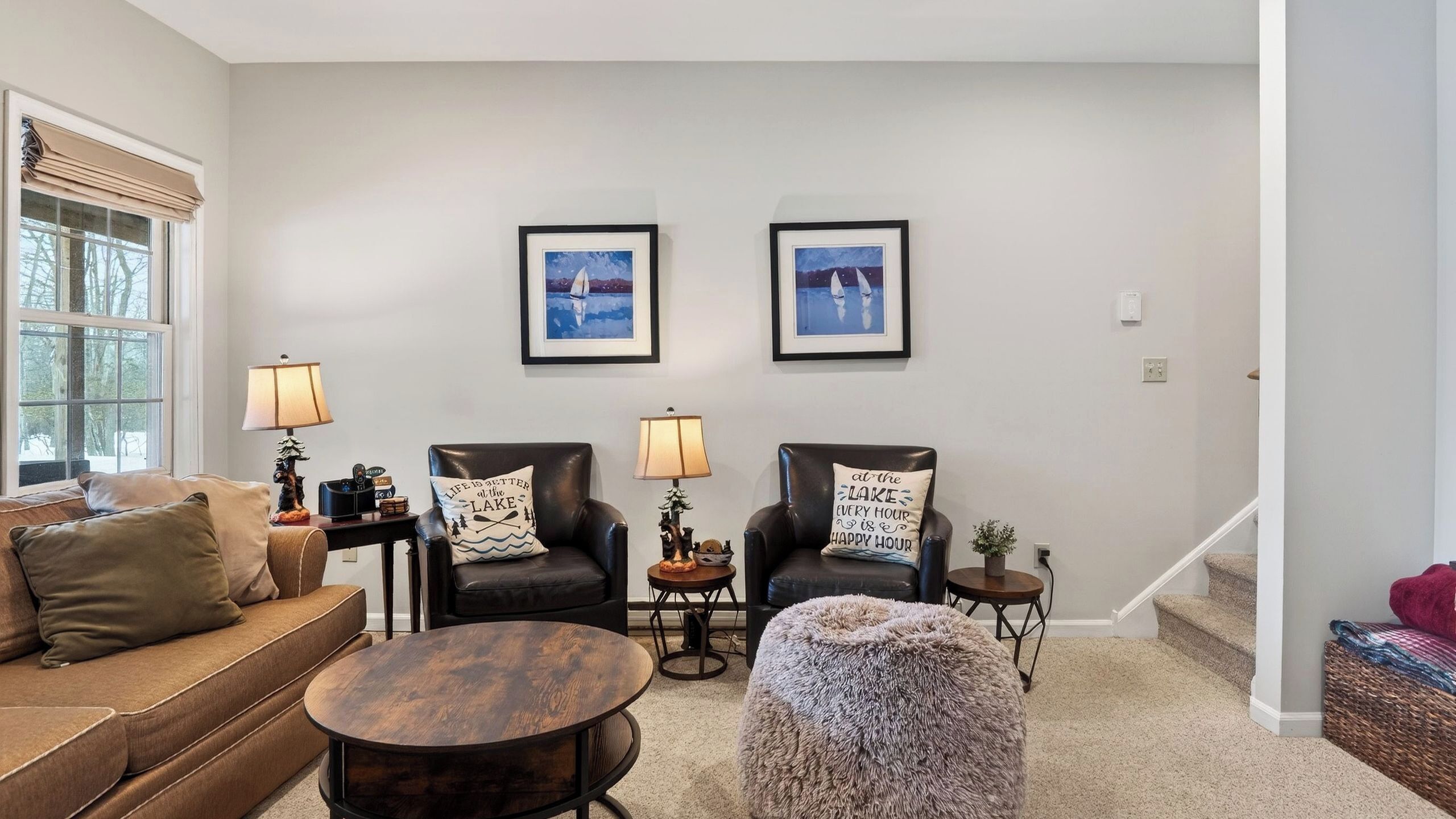 A living room with a sofa, two chairs, a round coffee table, and two lamps. There are framed pictures on the wall and a window with blinds.