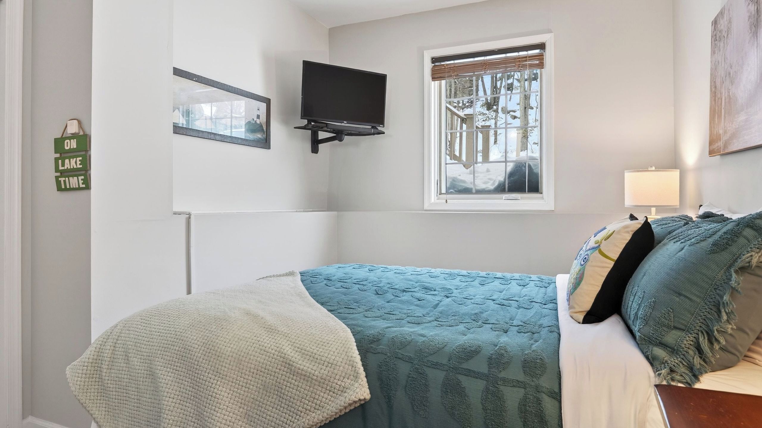 A bedroom with a bed, a flat-screen TV, a window with blinds, a wall-mounted mirror, and a sign on the wall.