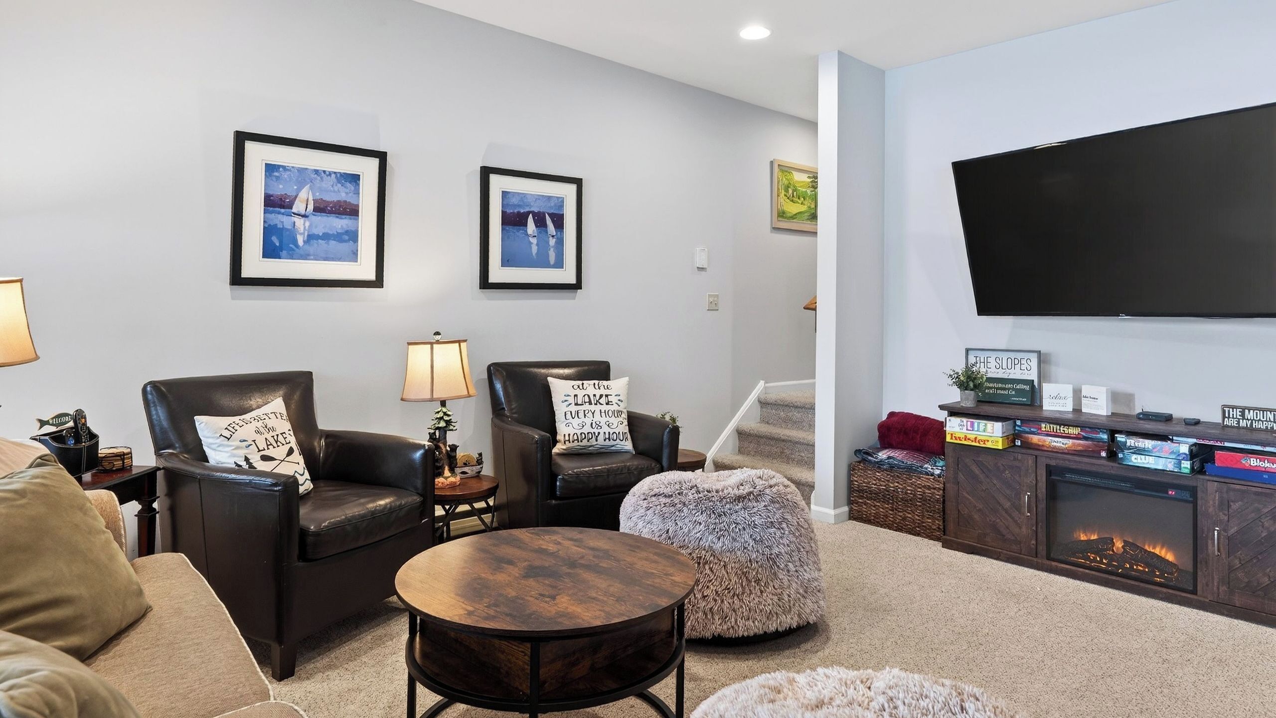 A modern living room with two leather armchairs, a round coffee table, a fireplace, and a flat-screen TV mounted on the wall.
