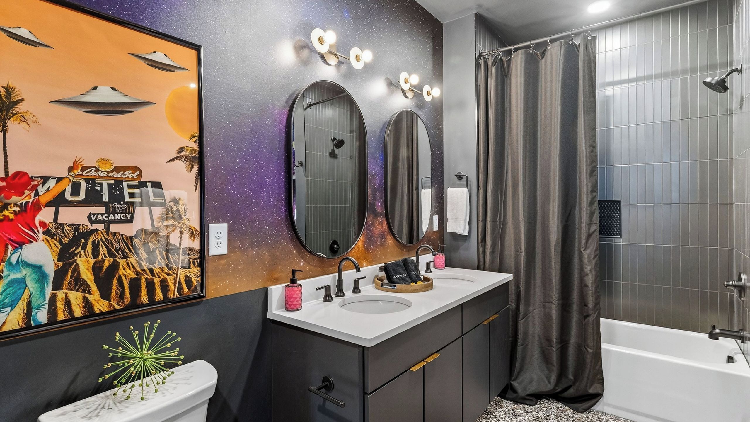 A modern bathroom with a large mirror, two sinks, a shower, and a wall with a space-themed mural.