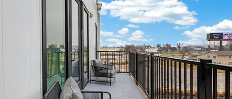 A balcony with a view of a parking lot and buildings, a chair, and a table.