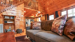L’intérieur d’un chalet accueillant, avec un escalier en bois, un canapé agrémenté de coussins à motifs, une bibliothèque et un coin cuisine.