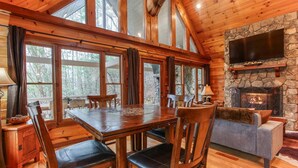 L’intérieur d’une cabane en rondins, avec une table à manger, des chaises, une cheminée et un téléviseur fixé à un mur en pierre.