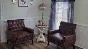 A room with two leather armchairs, a small round table with a lamp, framed floral artwork, and a large window with curtains. - Cozy 3-bedroom house in charming small town Sedley (Sedley)