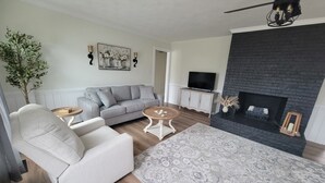 A living room with a grey sofa, a white armchair, a round coffee table, a fireplace, and a television. - Cozy 3-bedroom house in charming small town Sedley (Sedley)