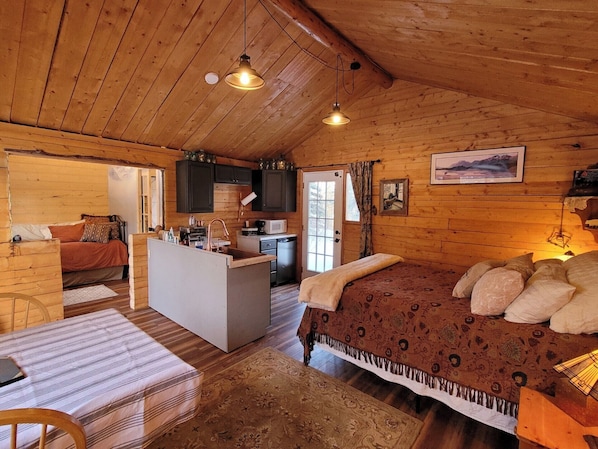 Interior acogedor de una cabaña con techo de madera, cama con ropa de cama estampada, zona de comedor y cocina con armarios oscuros.