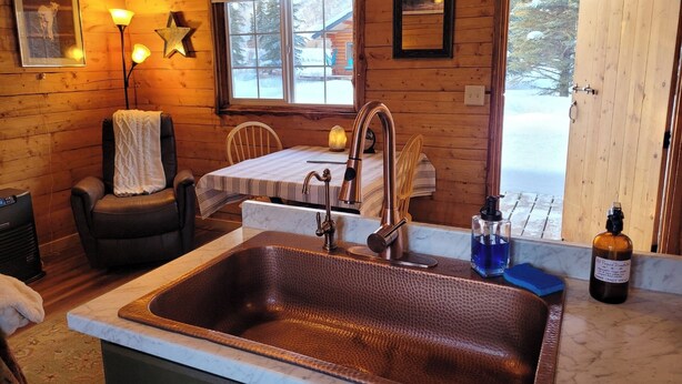 A cozy cabin interior with a wooden interior, a sink with a copper faucet, a chair with a towel, and a window with a view of a snowy landscape. - The Polaris is a cozy delight with 360 degree mountain views.  Pet Friendly! (Sutton)