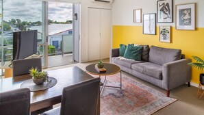 Ein modernes Wohnzimmer mit grauem Sofa, Couchtisch und Essbereich inklusive Tisch und Stühlen. Ein großes Fenster bietet Blick auf ein Gebäude und Bäume.