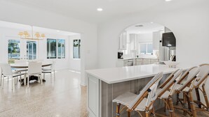 Una cuina moderna amb armaris blancs, una illa gran i una zona de dinar amb caires blancs i un llumser.