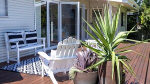Eine Terrasse mit weißen Stühlen und einem gemusterten Teppich, einer großen Kübelpflanze und einer Glastür zu einer überdachten Terrasse.
