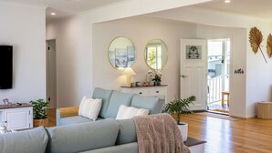 Ein Wohnzimmer mit grauem Sofa, einem weißen Schrank und einem Fernseher. An der Wand befinden sich zwei runde Spiegel, ein Deckenventilator und eine Tür, die auf einen Balkon führt.
