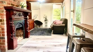 Una habitación con chimenea de ladrillo, techo de madera, ventana con vista a follaje, un sofá y una pequeña mesa con un ventilador.
