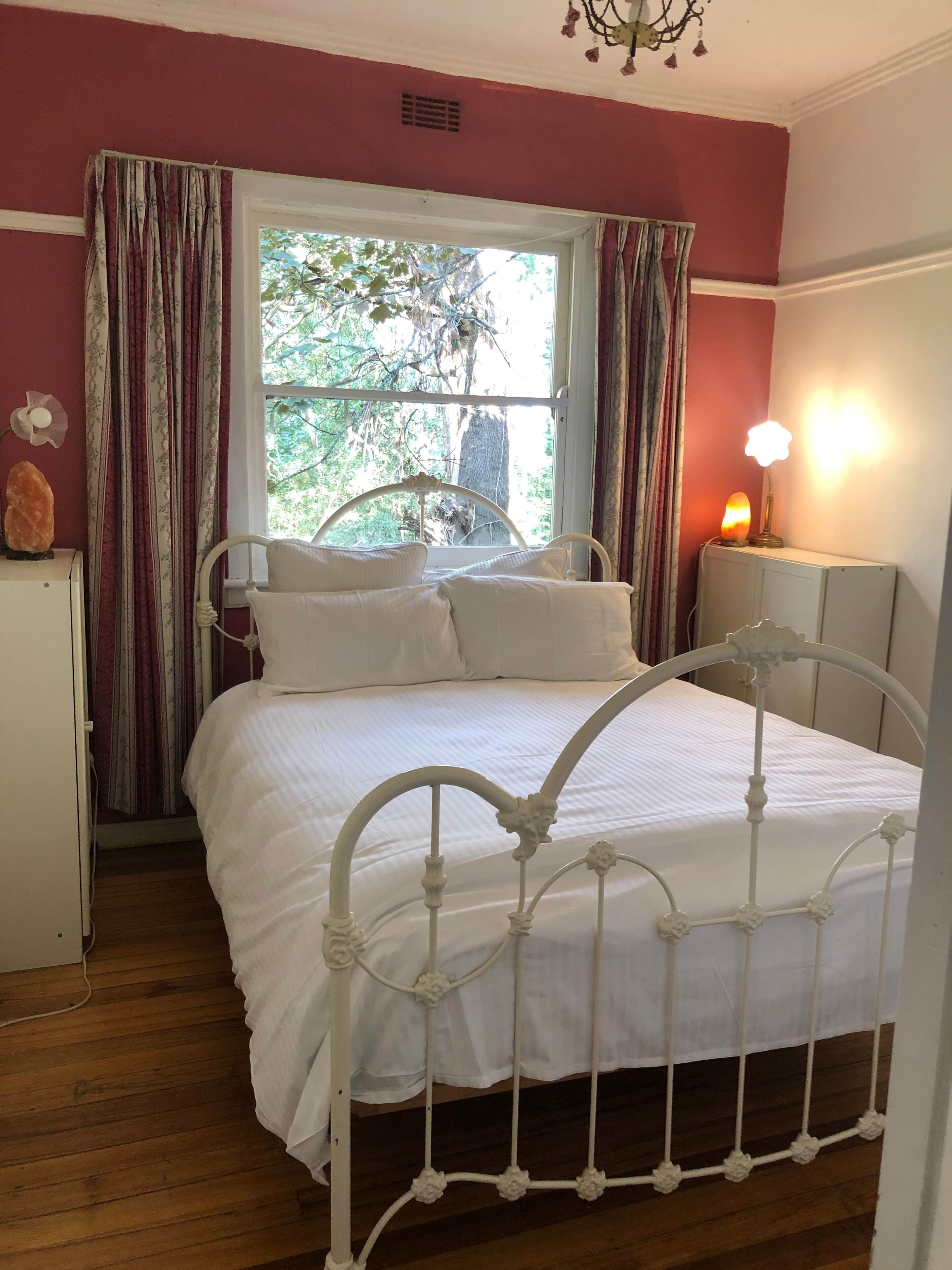 Een slaapkamer met een bed met een wit metalen frame, een bedlamp, een raam met gordijnen, en een kleine tafel met een bloem.