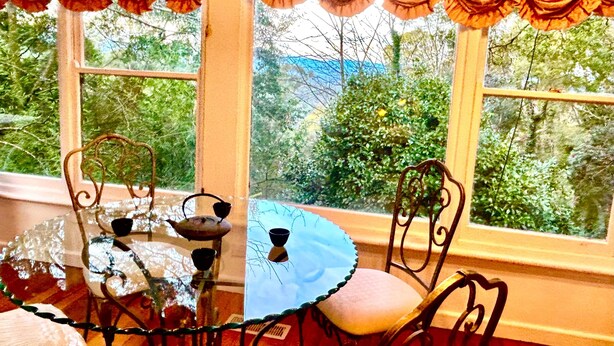 Un comedor con una mesa redonda y sillas, una ventana con cortinas y vistas al exterior a zonas verdes.