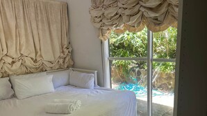 Una cama con ropa de cama blanca, una ventana con vista a follaje y una cortina.