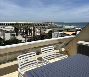 A balcony with white chairs and a table, overlooking a parking garage and a beach. - IRIS - Studio Héliopolis C13 - Luxury - 2 people (AGDE)
