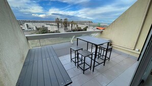 A balcony with a table and chairs, a view of buildings and palm trees, and a cloudy sky. - MEDUSE - Studio Héliopolis C29 - Luxury - 2 people (AGDE)